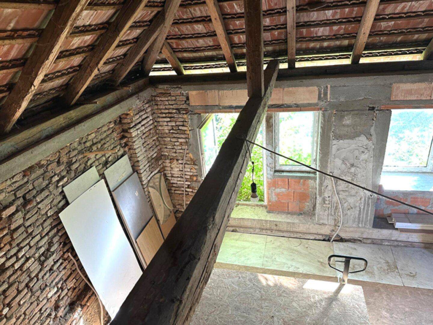 Dachbodenraum mit freiliegenden Ziegelwänden, Holzbalken und einem Fenster mit Blick ins Grüne.