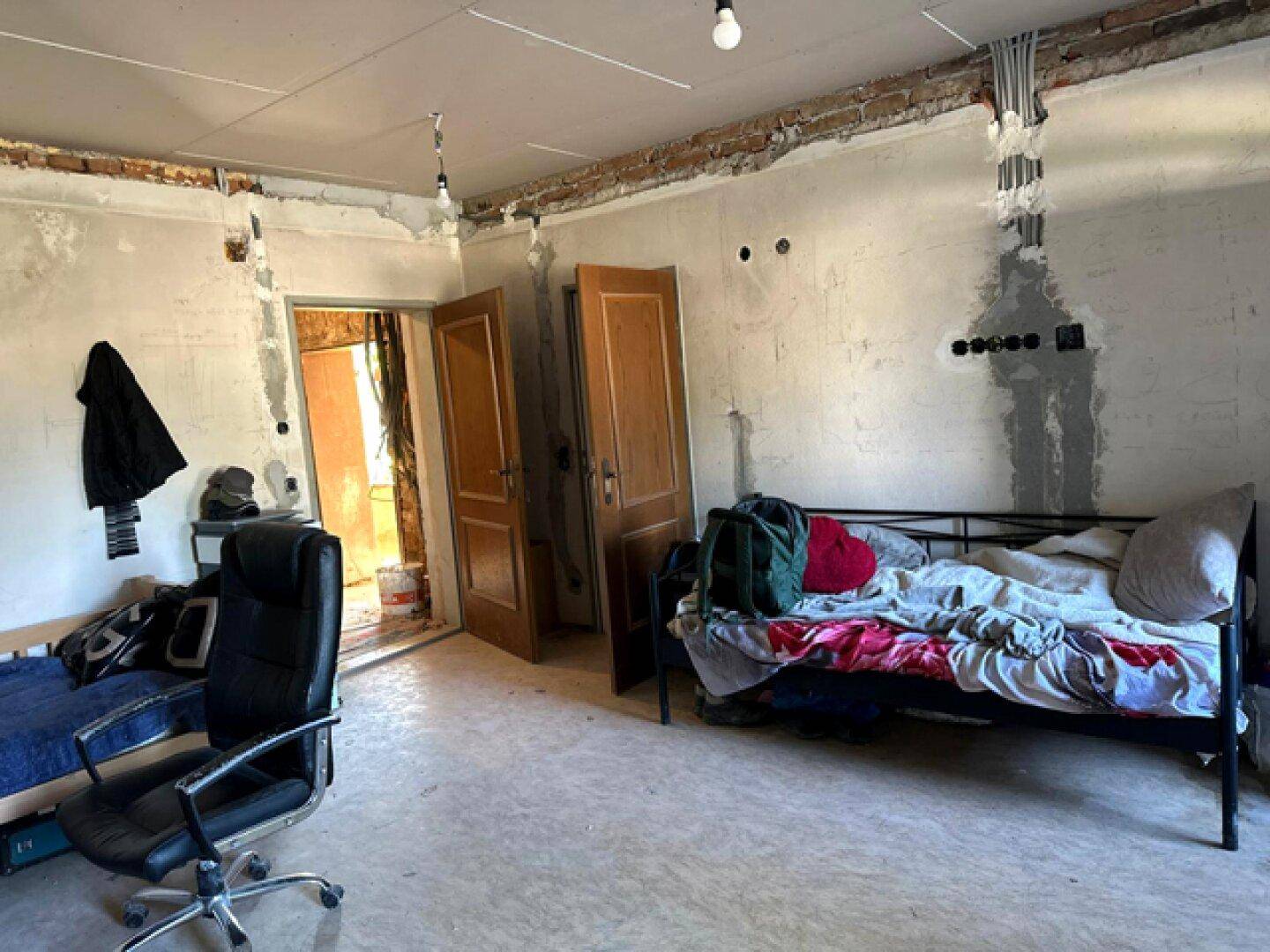 Schlafzimmer im Rohbau mit Bett, Schreibtisch und sichtbaren Installationen.