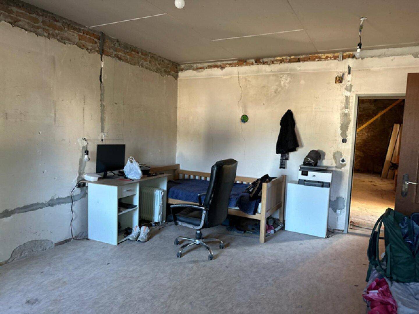 Einfaches Schlafzimmer im Rohbau mit Bett und Arbeitsbereich, bereit zur Renovierung.