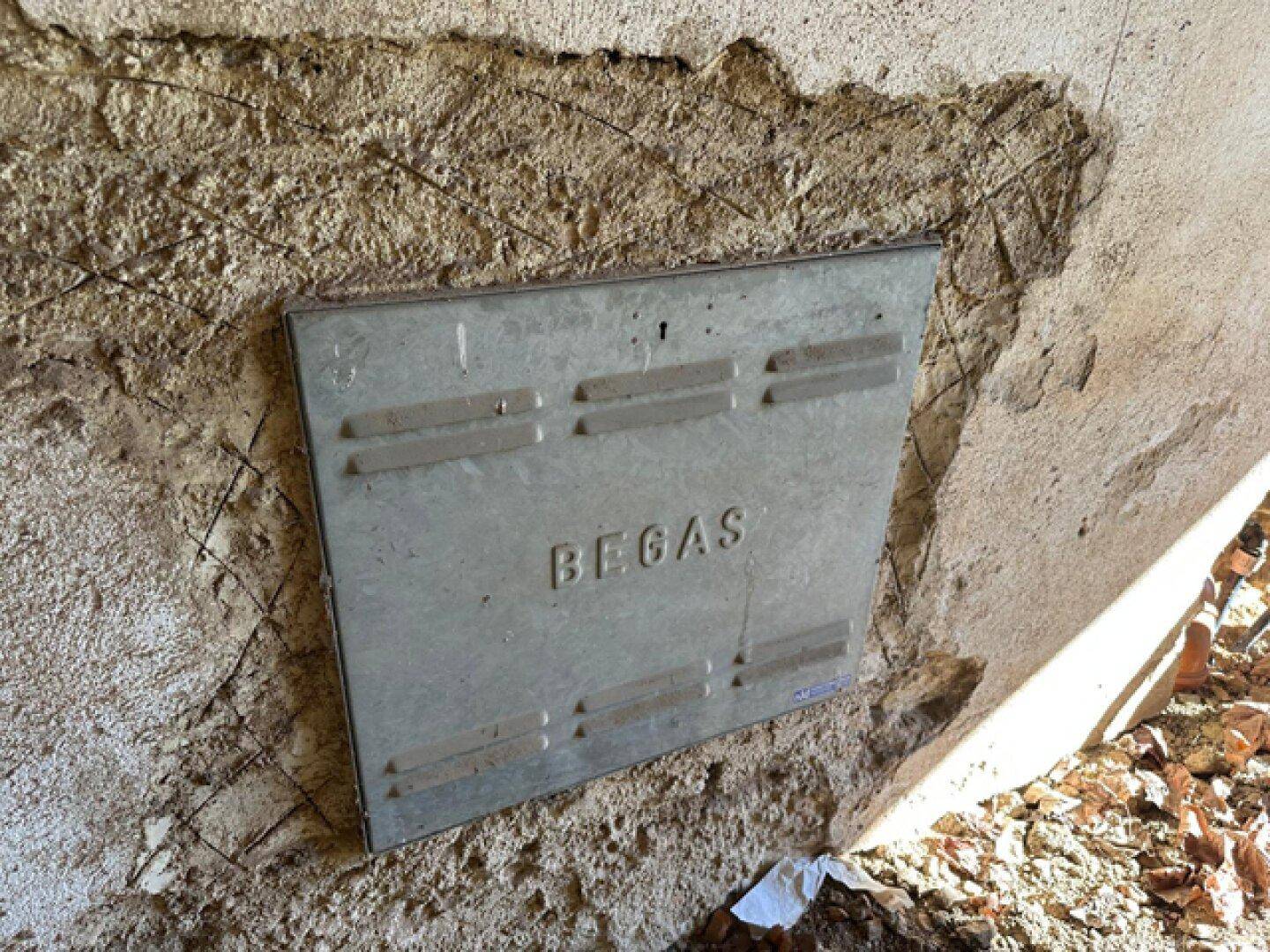 Metallplatte mit der Aufschrift "BEGAS" an einer rauen, beschädigten Außenwand des Gebäudes.