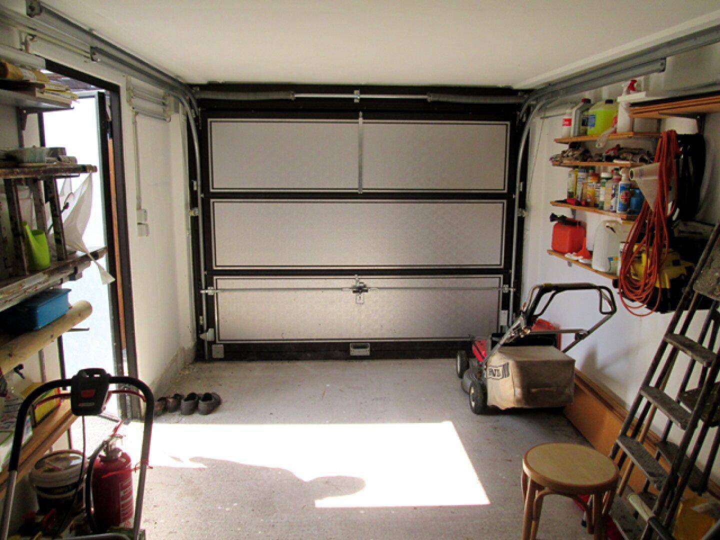 Innenansicht einer Garage mit Rolltor, Rasenmäher und Regalen für Werkzeuge.
