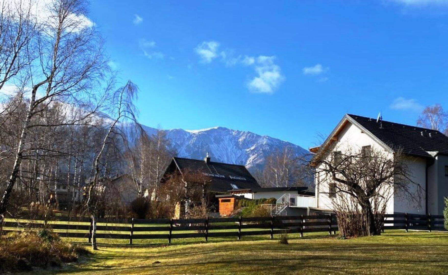 Häuser in ländlicher Umgebung mit schneebedeckten Bergen im Hintergrund unter blauem Himmel.