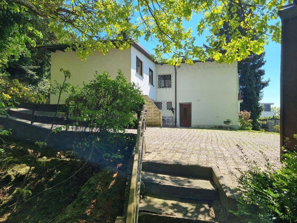 Weißes Wohnhaus mit gepflastertem Vorplatz, umgeben von hohen Bäumen und viel Grün.