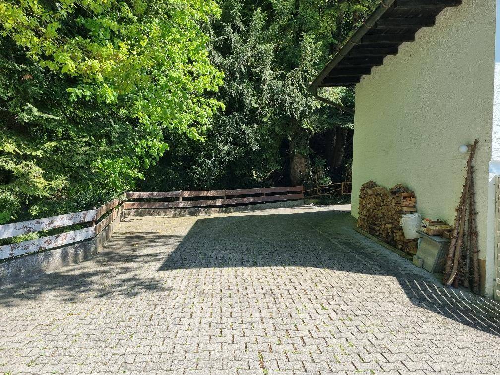 Gepflasterter Parkplatz oder Zufahrt neben dem Haus, umgeben von Bäumen und gestapeltem Brennholz.