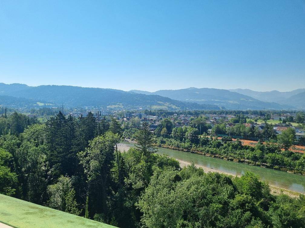 Weitläufiger Blick über grüne Wälder, einen Fluss und eine Stadtlandschaft mit Bergen im Hintergrund.