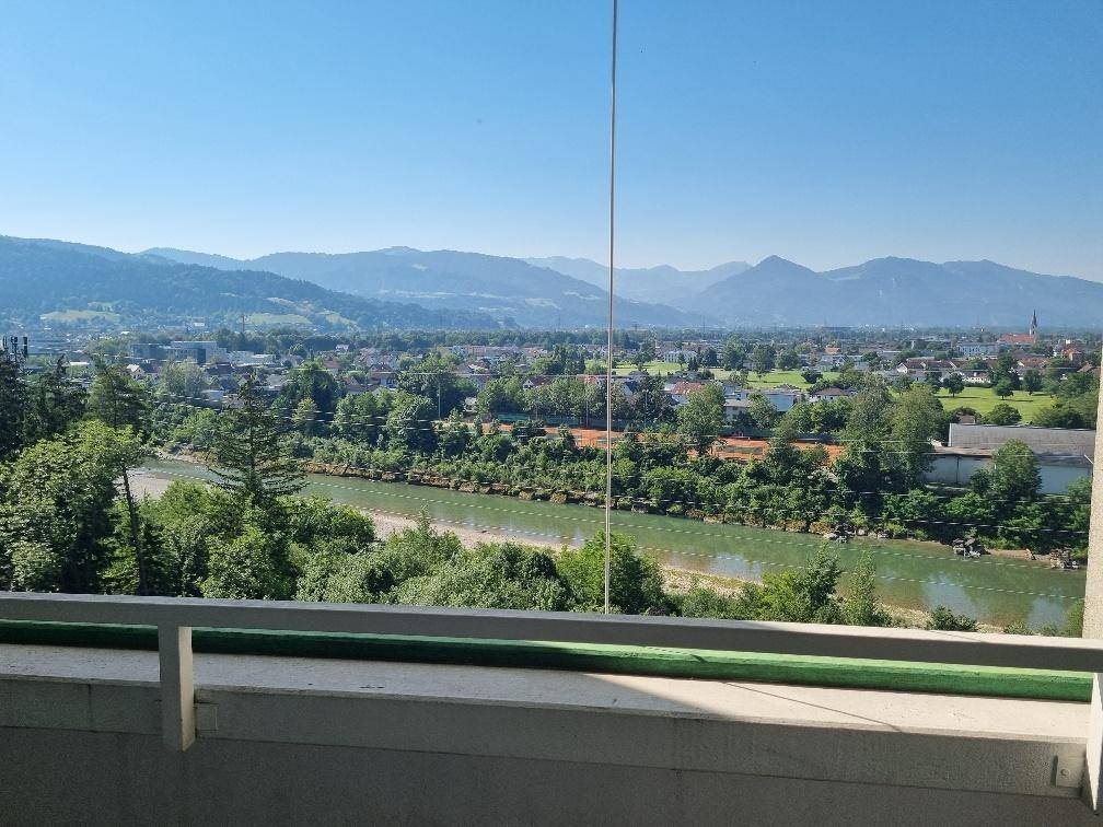 Balkon mit Geländer und weitem Blick auf einen Fluss, grüne Bäume und Berge.