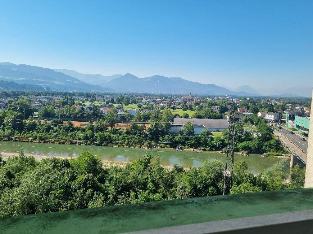 Panoramablick von einem Balkon auf einen Fluss, Tennisplätze und eine Stadt vor Bergkulisse.