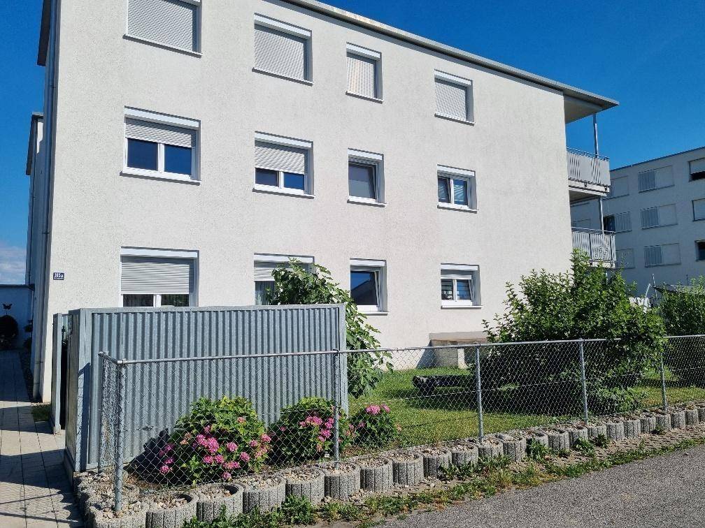 Seitenansicht eines Mehrfamilienhauses mit Rollläden und einem kleinen eingezäunten Gartenbereich.