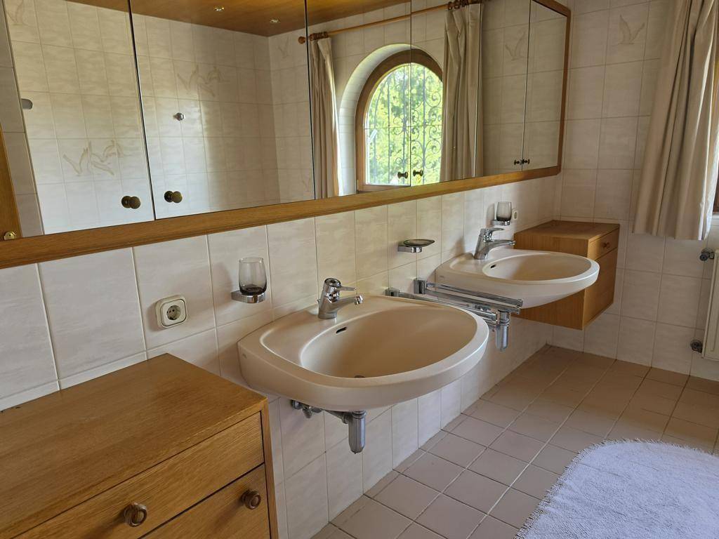 Helles Badezimmer mit Doppelwaschbecken, Spiegelschrank und beigefarbenen Fliesen an Wänden und Boden.