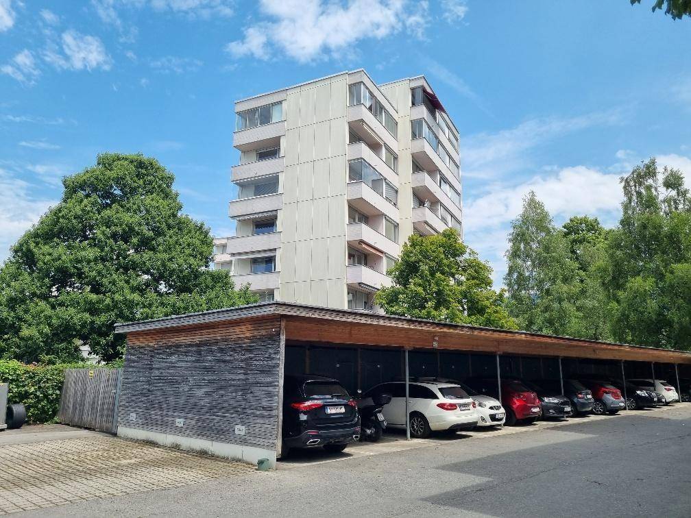 Wohngebäude mit Carport-Anlage im Vordergrund, umgeben von grünen Bäumen.