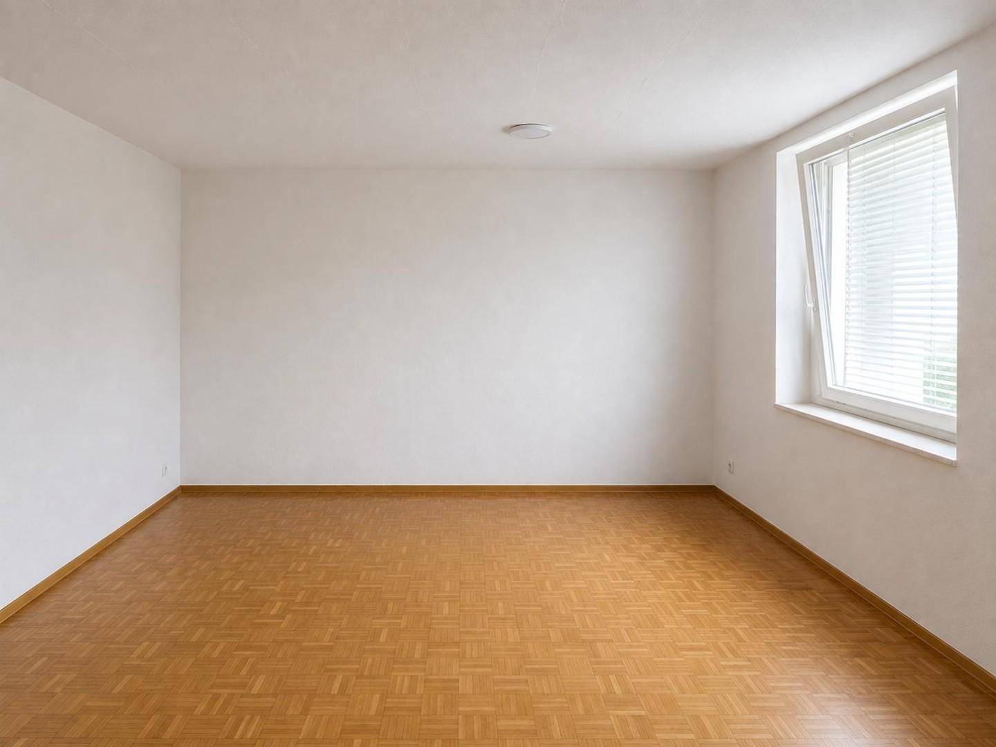 Heller, leerer Raum mit Parkettboden und Fenster mit Jalousien, bietet viel Tageslicht.