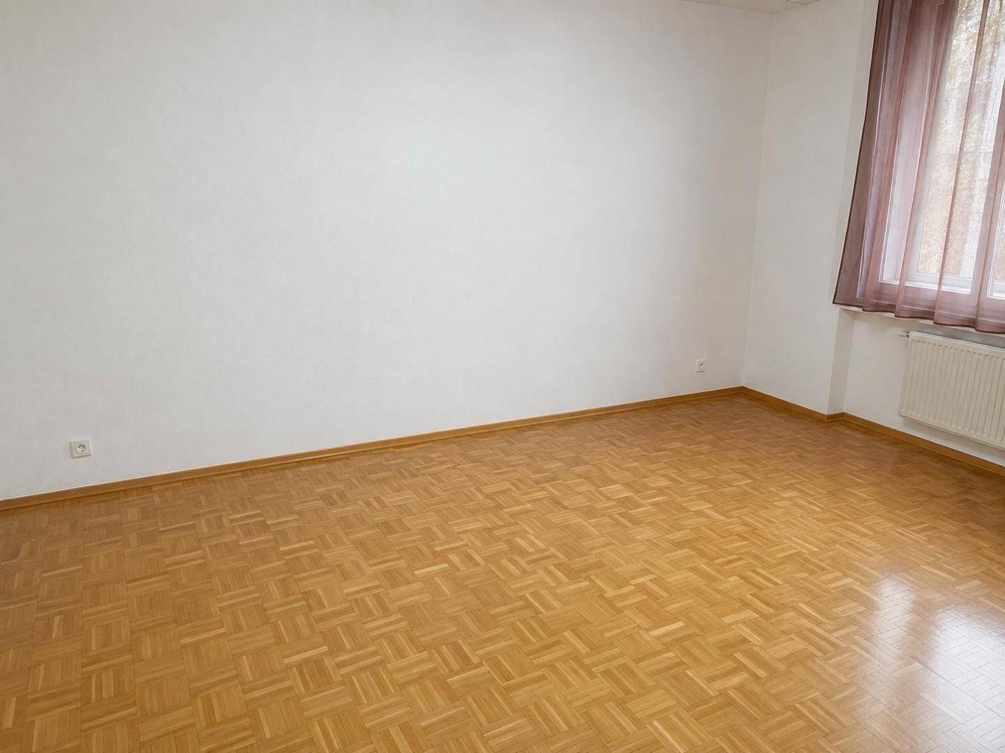 Leerer Raum mit hellem Parkettboden und Fenster mit dunklen Vorhängen, bereit zur Einrichtung.