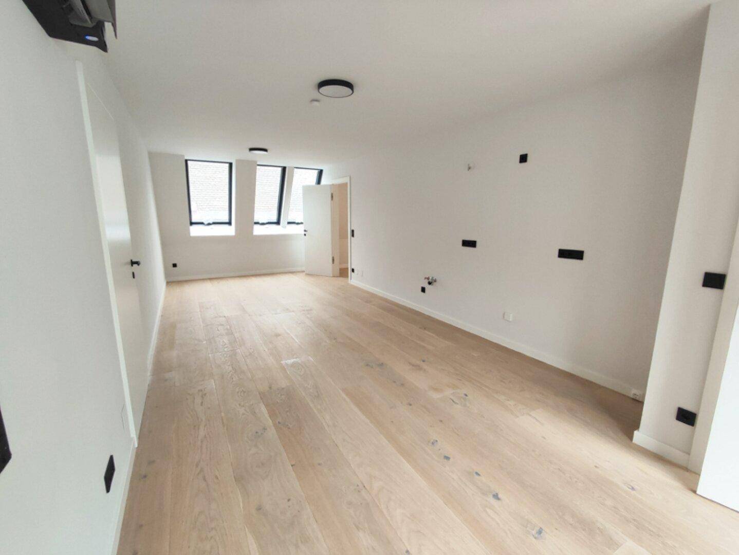 Geräumiger, heller Wohnbereich mit Holzboden und mehreren Fenstern, die viel Tageslicht hereinlassen.