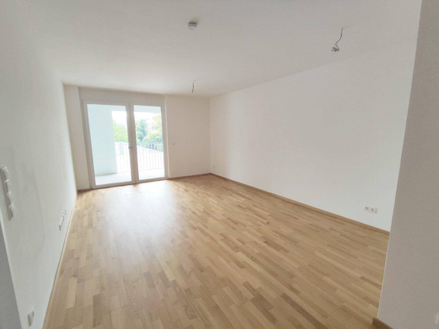 Heller, leerer Wohnraum mit hellem Parkettboden und Zugang zu einem Balkon.