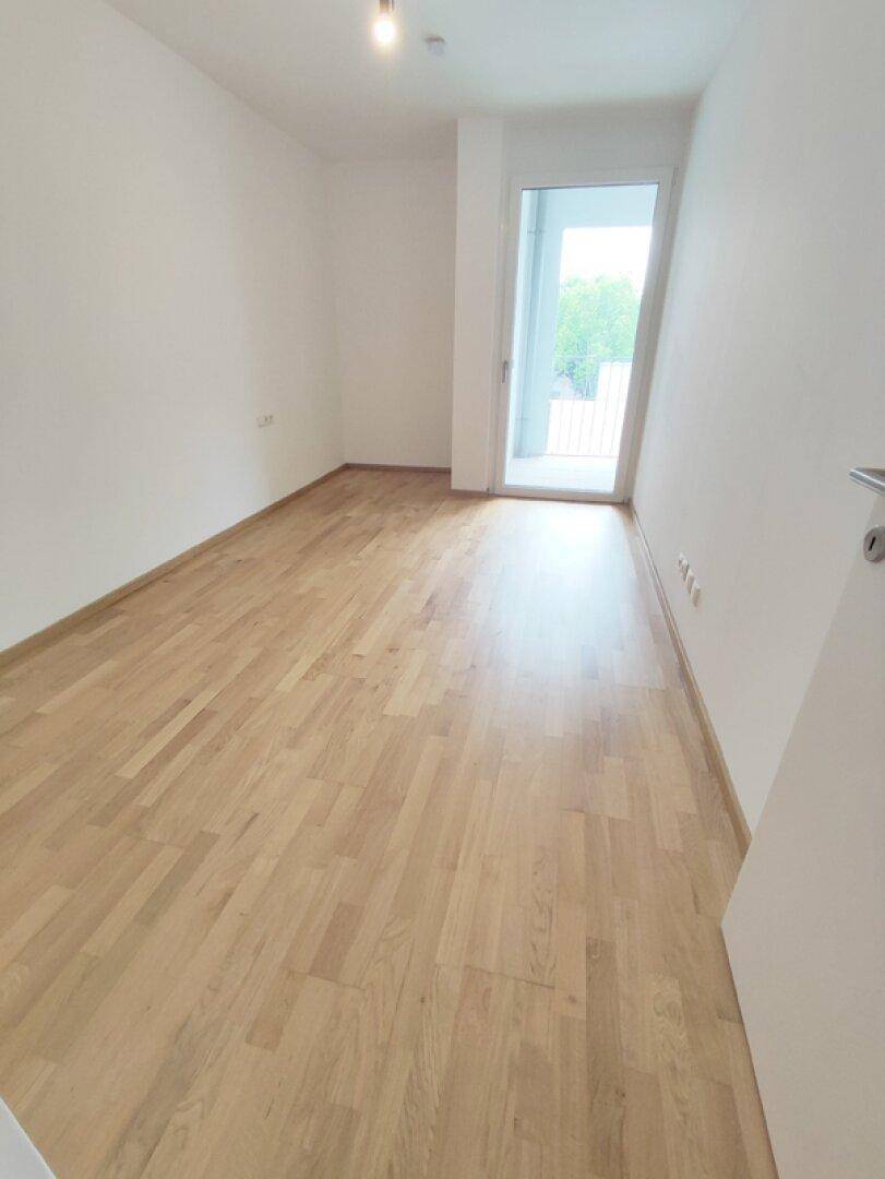 Heller, leerer Raum mit Parkettboden und Zugang zu einem französischen Balkon oder Fenster.