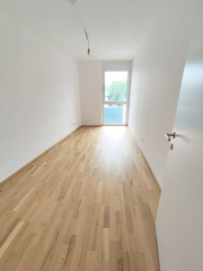Unmöblierter Raum mit hellem Parkettboden und einem großen Fenster, das viel Licht hereinlässt.