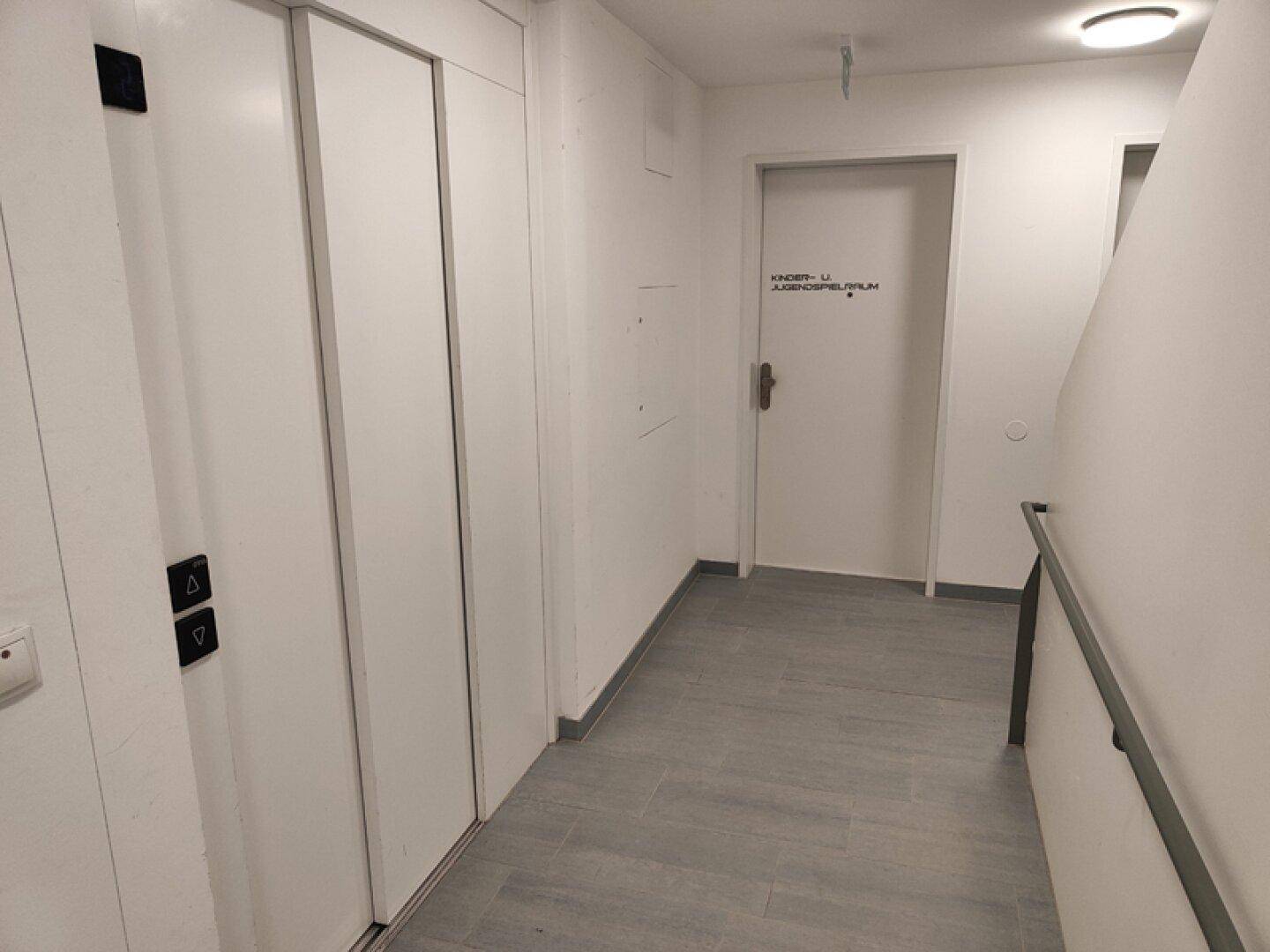 Heller Flur mit Aufzug, Zugang zum Kinderspielraum und Handlauf an der Wand.