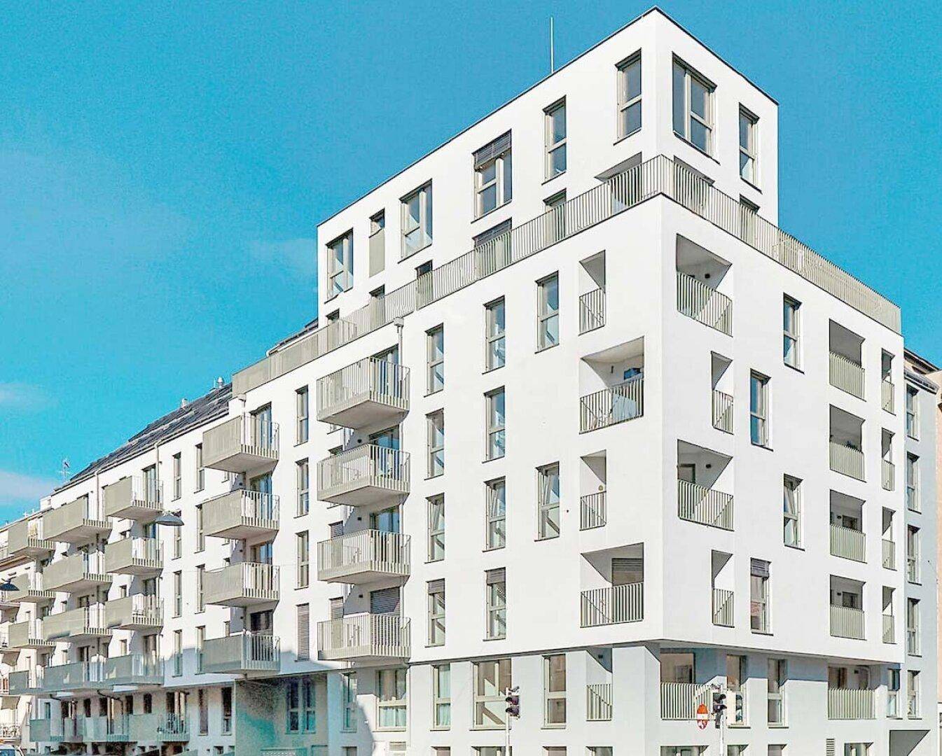 Modernes weißes Wohngebäude mit vielen Balkonen unter klarem blauem Himmel.