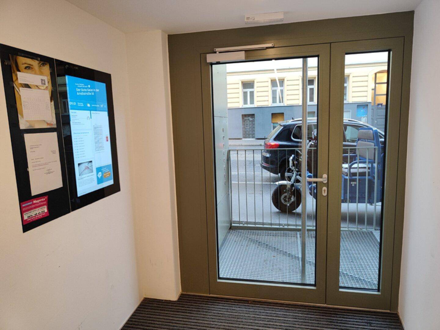 Eingangsbereich mit Glastür zur Straße und digitaler Anzeige an der Wand.