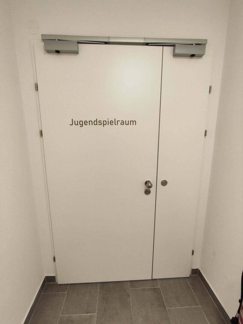 Weiße Doppeltür mit der Aufschrift "Jugendspielraum" in einem Gemeinschaftsbereich des Gebäudes.
