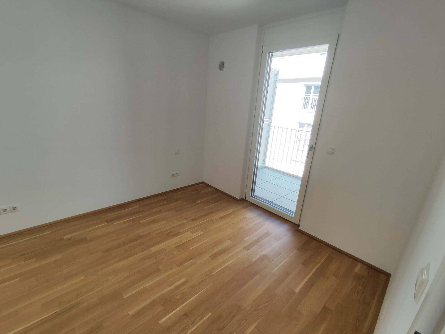 Unmöbliertes Schlafzimmer mit hellem Parkettboden und direktem Zugang zu einem Balkon.