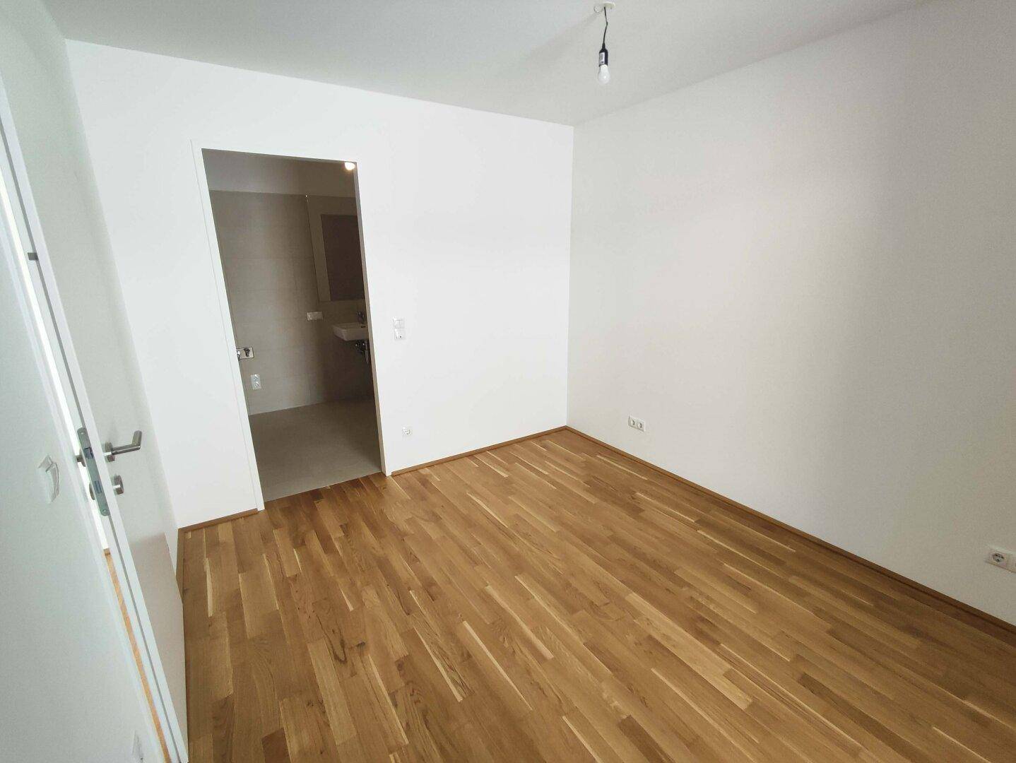 Leeres Schlafzimmer mit hellem Parkettboden und Zugang zu einem angrenzenden Badezimmer.