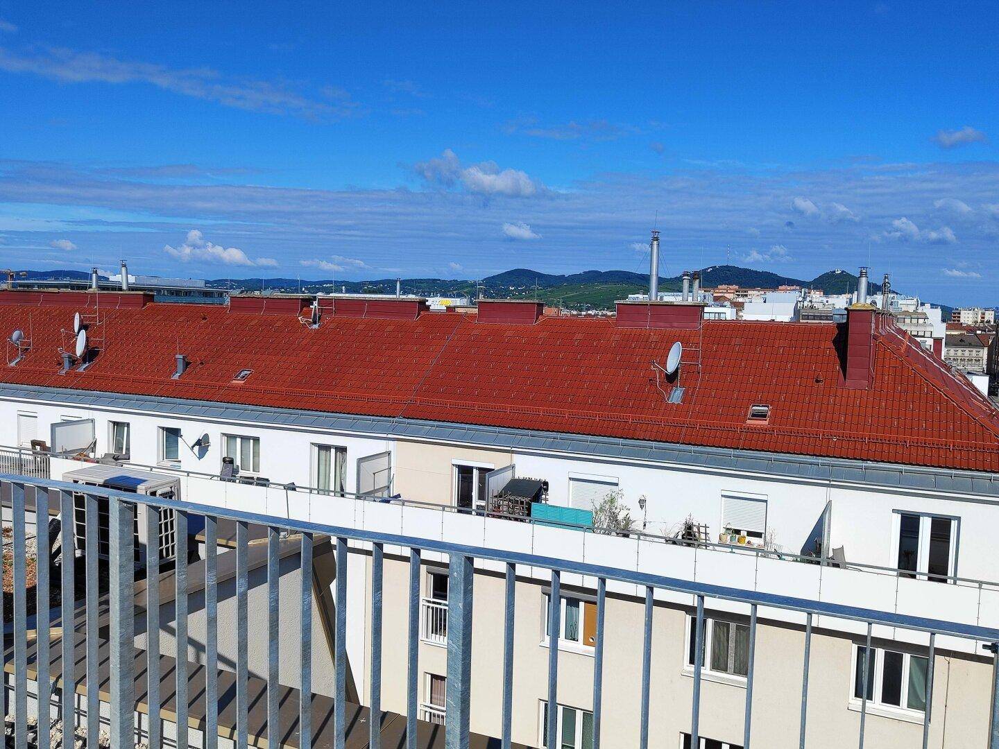 Weitläufiger Blick über die Dächer der Stadt mit grünen Hügeln am Horizont unter blauem Himmel.