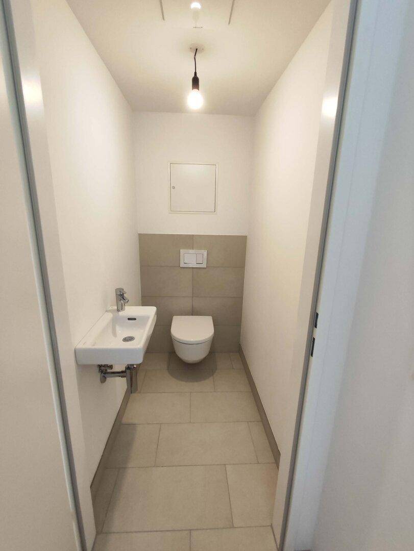 Modernes Gäste-WC mit Wand-WC, kleinem Waschbecken und hellen Fliesen.