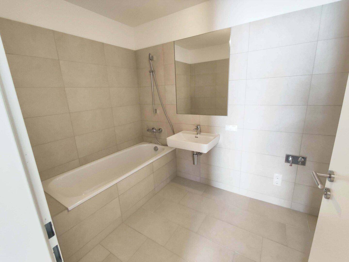 Helles Badezimmer mit Badewanne, Wandwaschbecken und großen beigefarbenen Fliesen.
