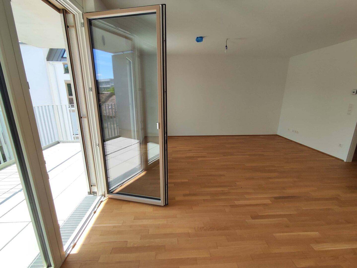 Heller Raum mit Holzboden und Zugang zu einem Balkon, der viel Tageslicht hereinlässt.