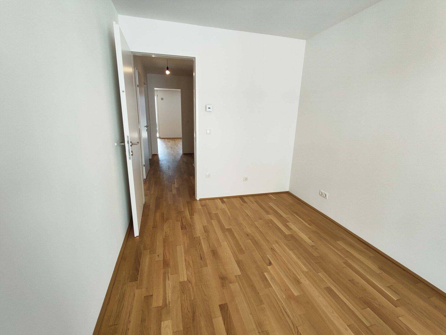 Heller, leerer Raum mit Parkettboden und Zugang zu einem langen Flur, ideal als Schlafzimmer.