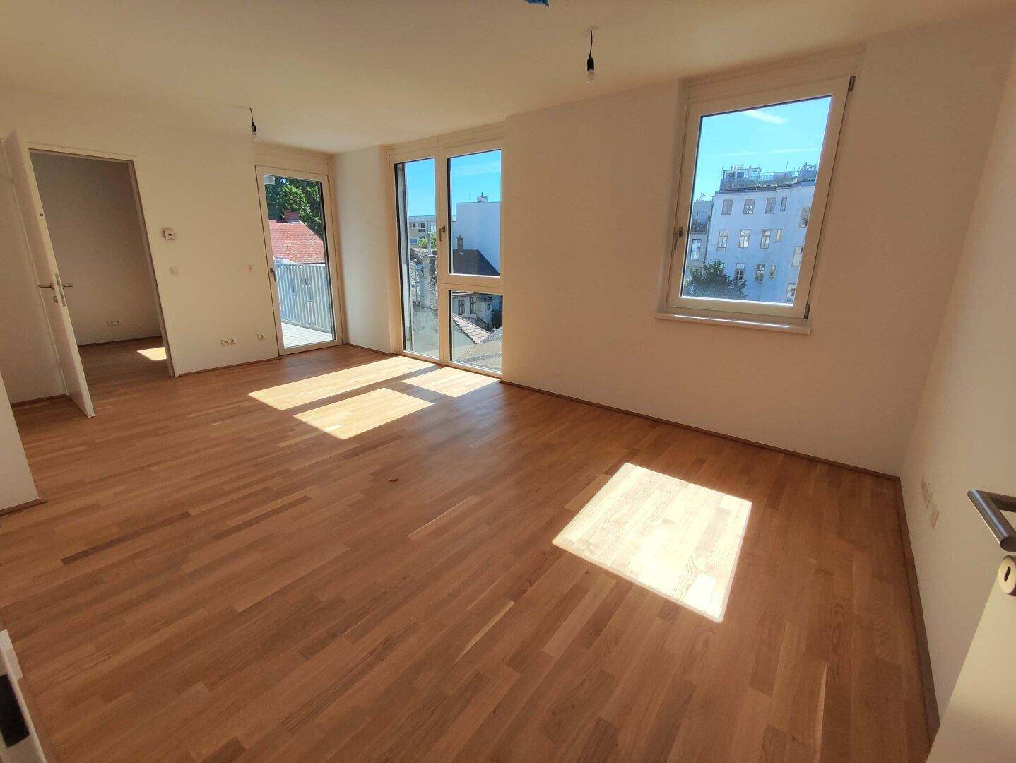 Heller, leerer Wohnraum mit Eichenparkettboden, großen Fenstern und Zugang zu einem Balkon.