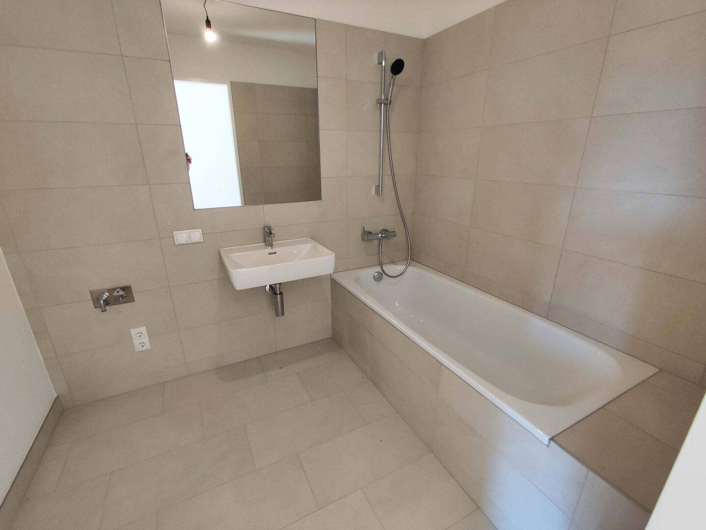 Modernes Badezimmer mit hellgrauen Fliesen, einer Badewanne mit Duschfunktion und einem Einzelwaschbecken.