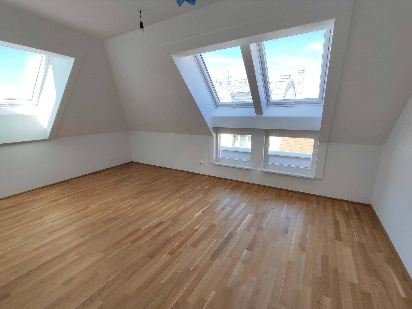 Helles Zimmer im Dachgeschoss mit Holzboden, schrägen Wänden und mehreren Dachfenstern für viel Licht.