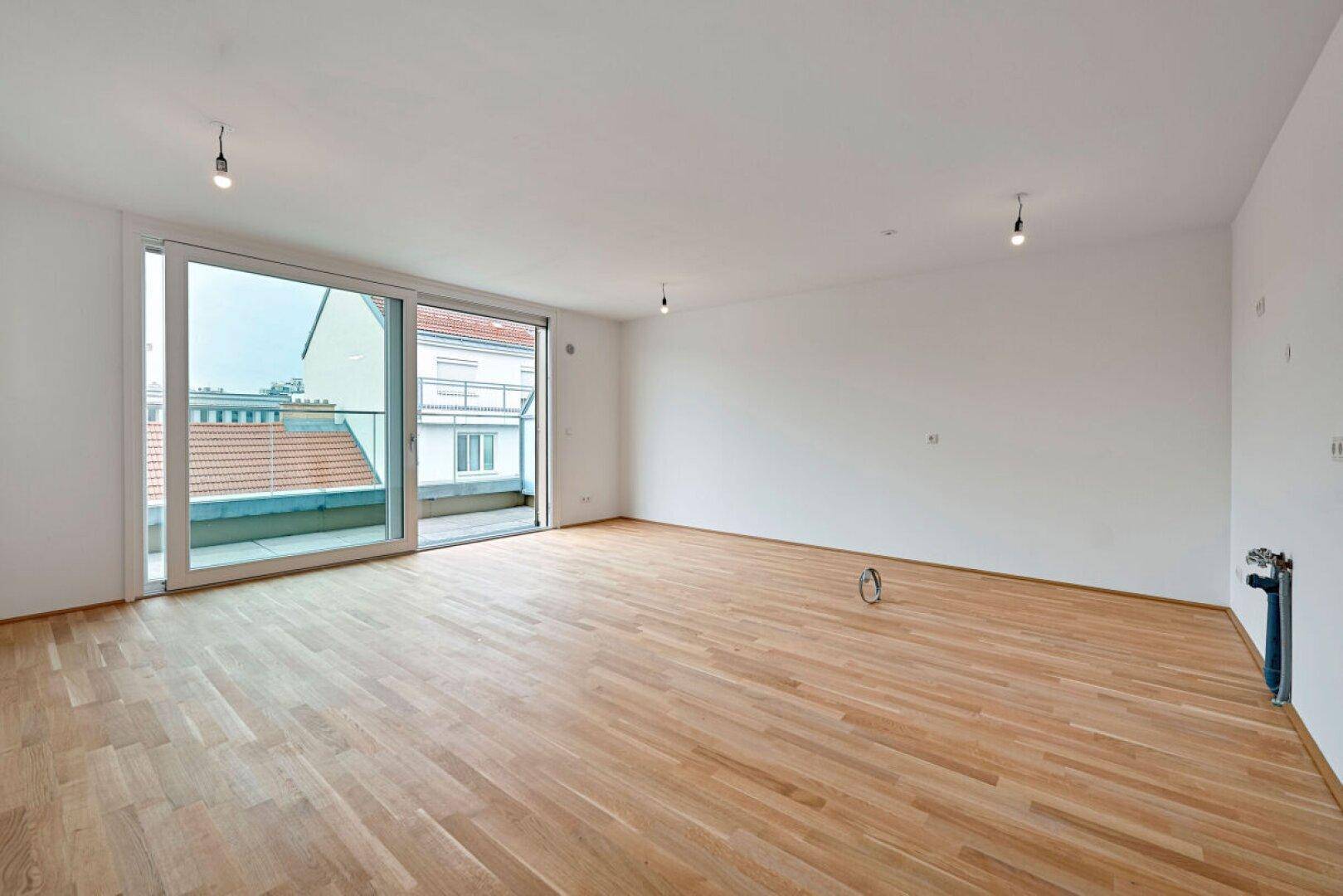 Geräumiger, leerer Raum mit hellem Holzboden und Zugang zu einem Balkon, ideal für einen Wohnbereich.