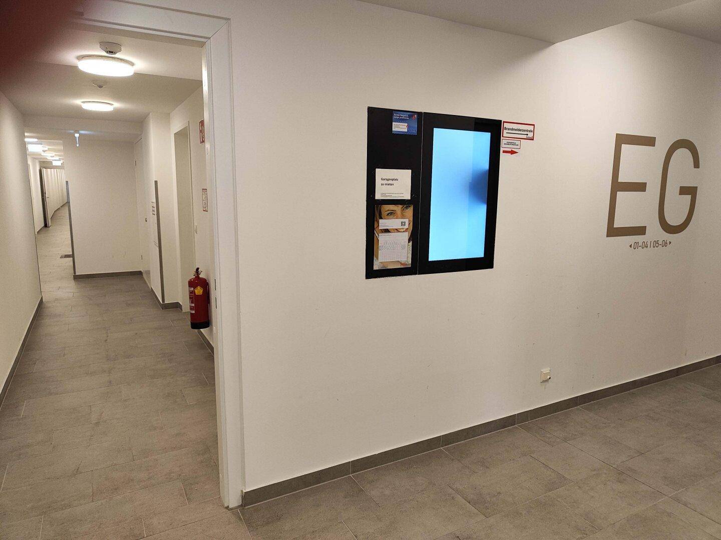 Heller Gemeinschaftsflur im Erdgeschoss mit Fliesenboden und Informationsanzeige an der Wand.