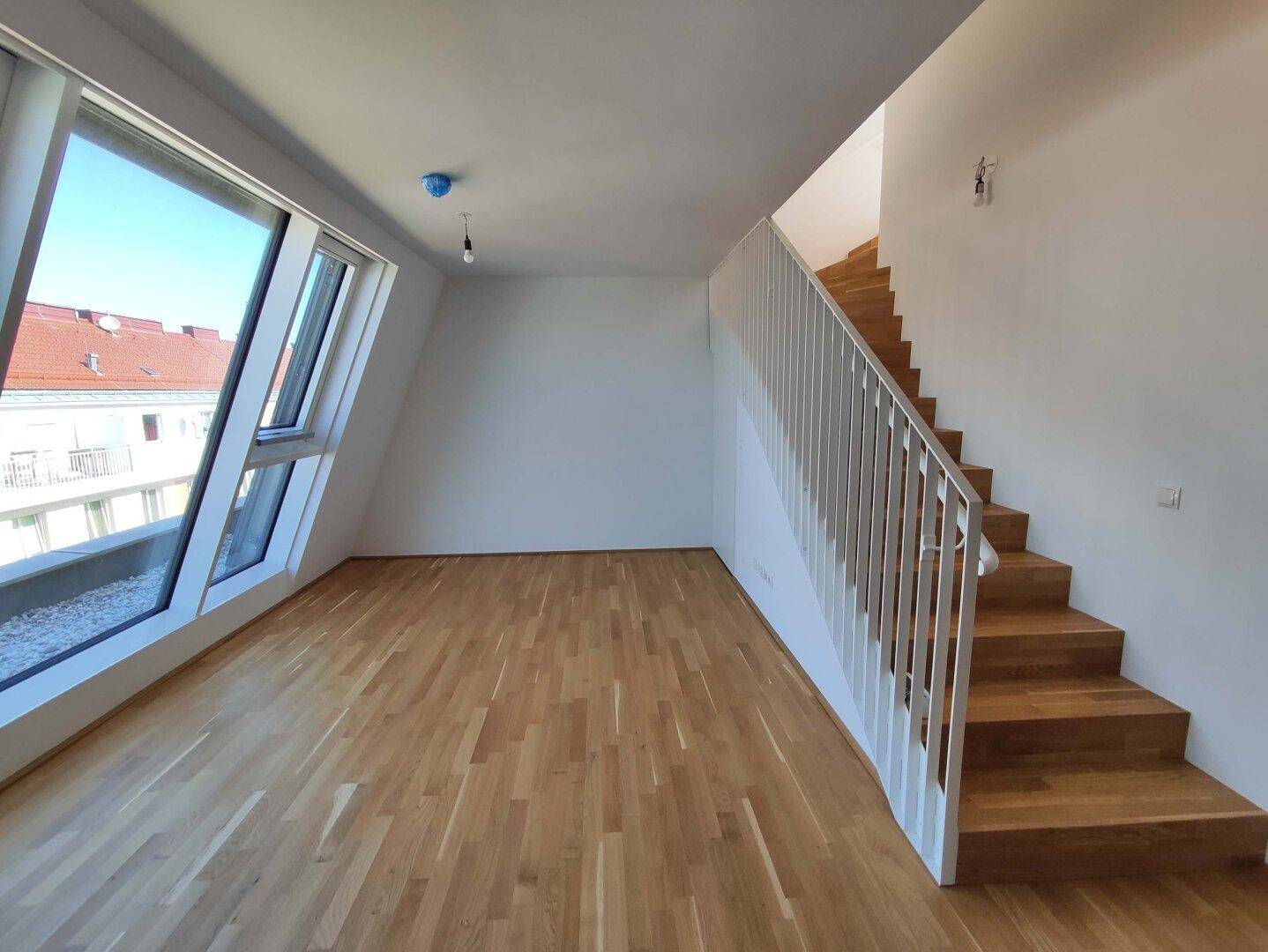 Geräumiger Wohnbereich mit Holztreppe, Parkettboden und großen Fenstern, die viel Licht hereinlassen.