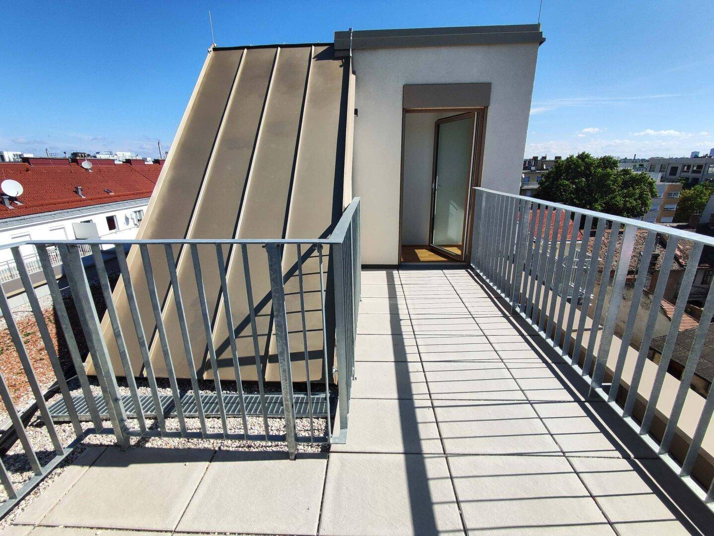 Geräumige Dachterrasse mit Metallgeländer und Blick auf die umliegenden Gebäude unter blauem Himmel.