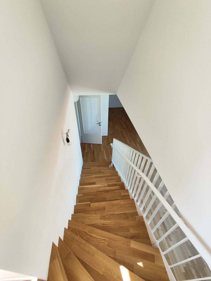 Holztreppe mit weißem Geländer führt in einen hellen Flur mit Parkettboden und weißen Wänden.