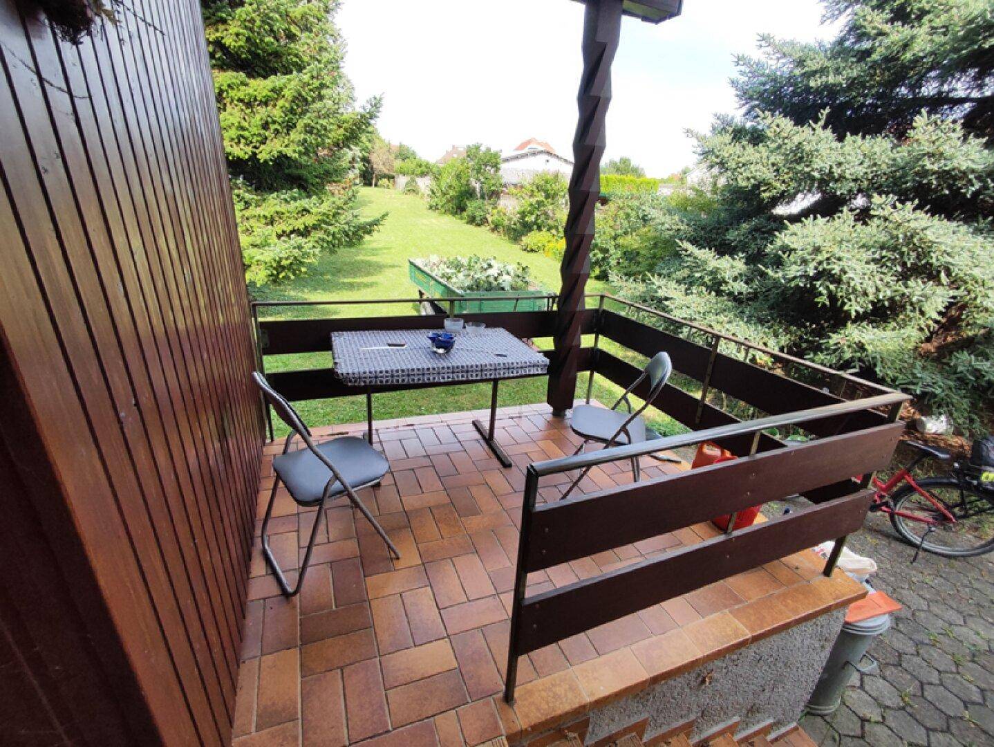 Kleiner überdachter Balkon mit Fliesenboden und einfacher Sitzgelegenheit, umgeben von Bäumen.