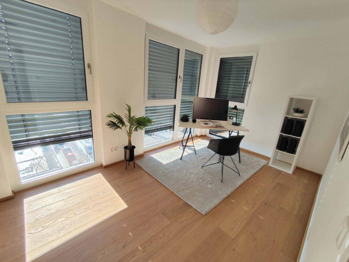 Helles Arbeitszimmer mit Schreibtisch, Holzboden und großen Fenstern mit Jalousien.