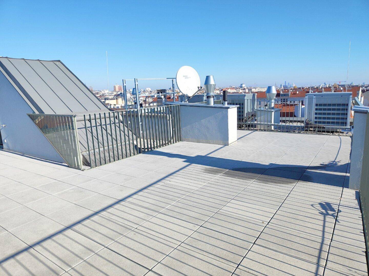 Weitläufige Dachterrasse mit Fliesen, Metallgeländer und Blick auf die Stadtsilhouette.