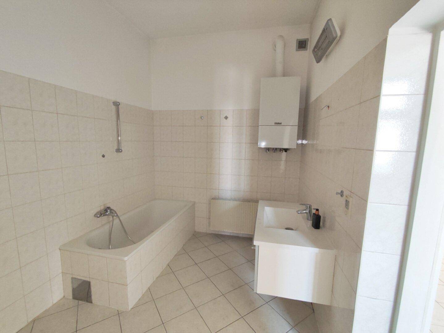 Gefliestes Badezimmer mit Badewanne, Waschbecken und einem an der Wand montierten Warmwasserbereiter.