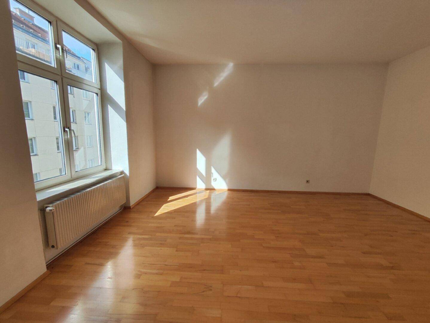 Lichtdurchflutetes Zimmer mit glänzendem Parkettboden und einem großen Fenster, das viel Sonne hereinlässt.