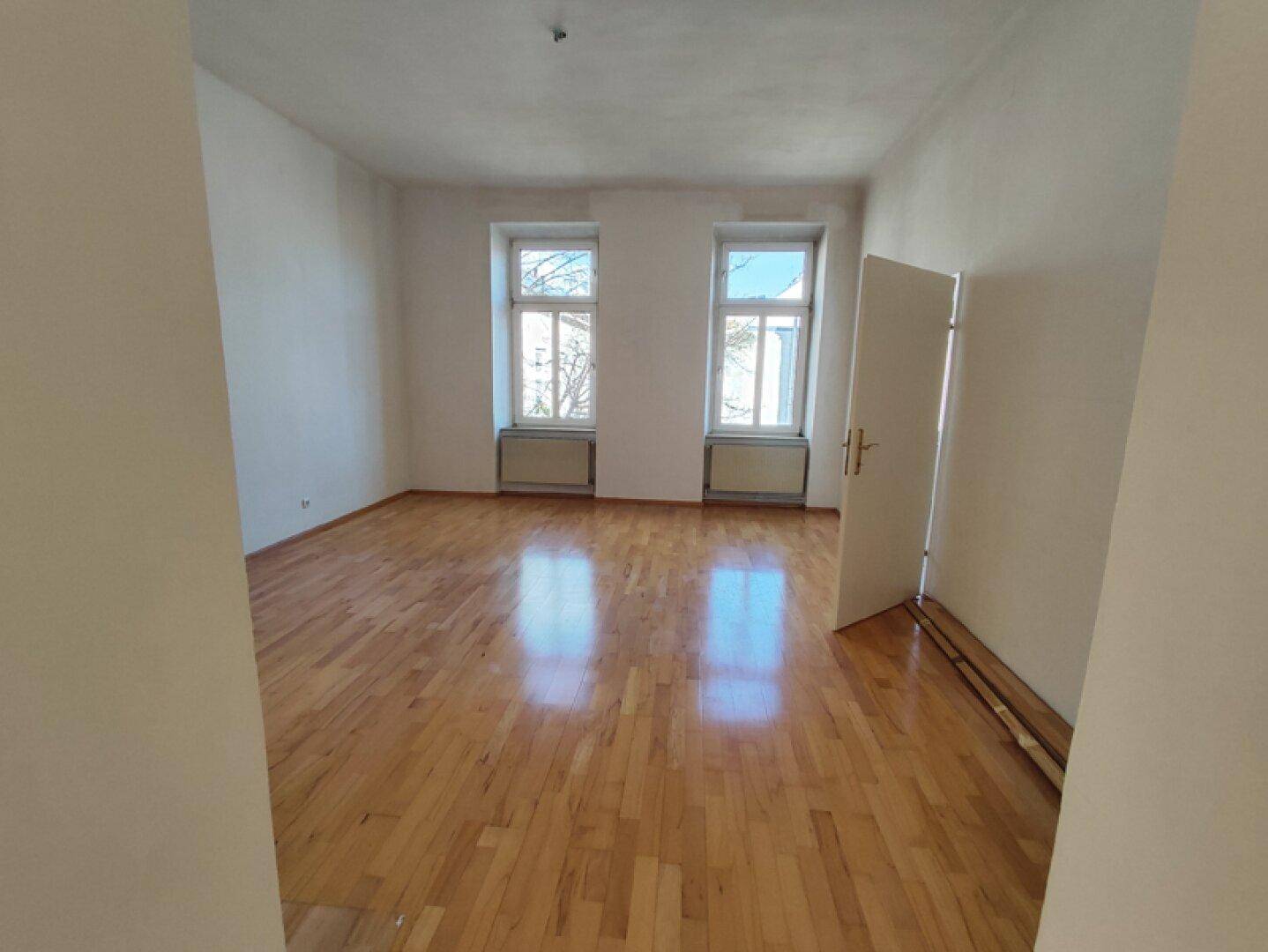 Helles Zimmer mit Parkettboden und zwei Fenstern, die einen Blick ins Grüne bieten.