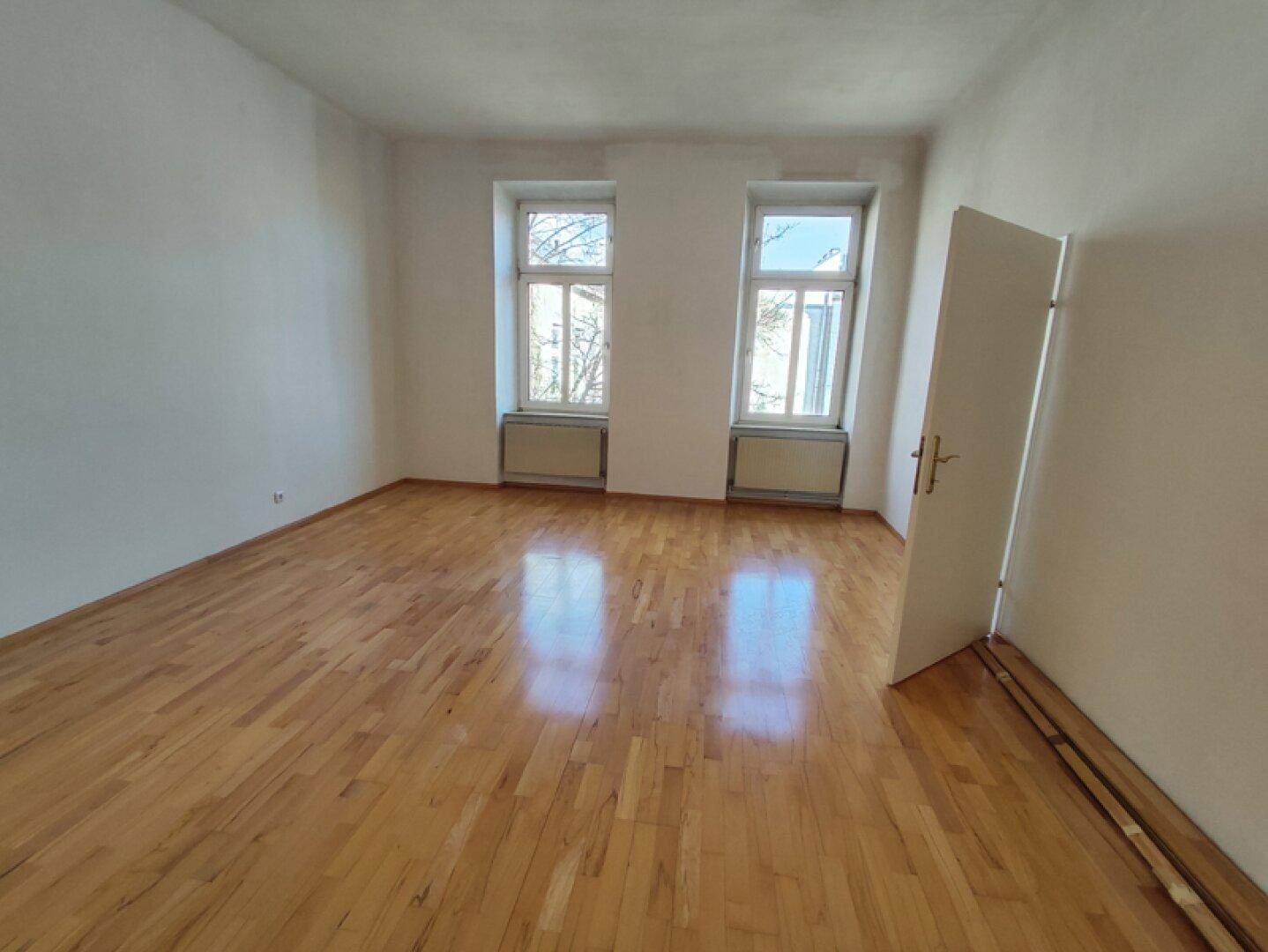 Geräumiges Zimmer mit glänzendem Parkettboden und Blick auf die Bäume vor dem Fenster.