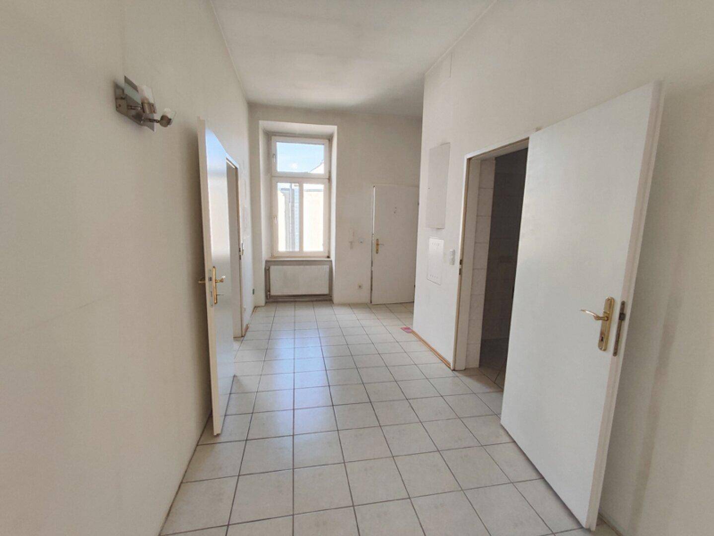Langer Flur mit Fliesenboden und einem Fenster am Ende, das Tageslicht hereinlässt.