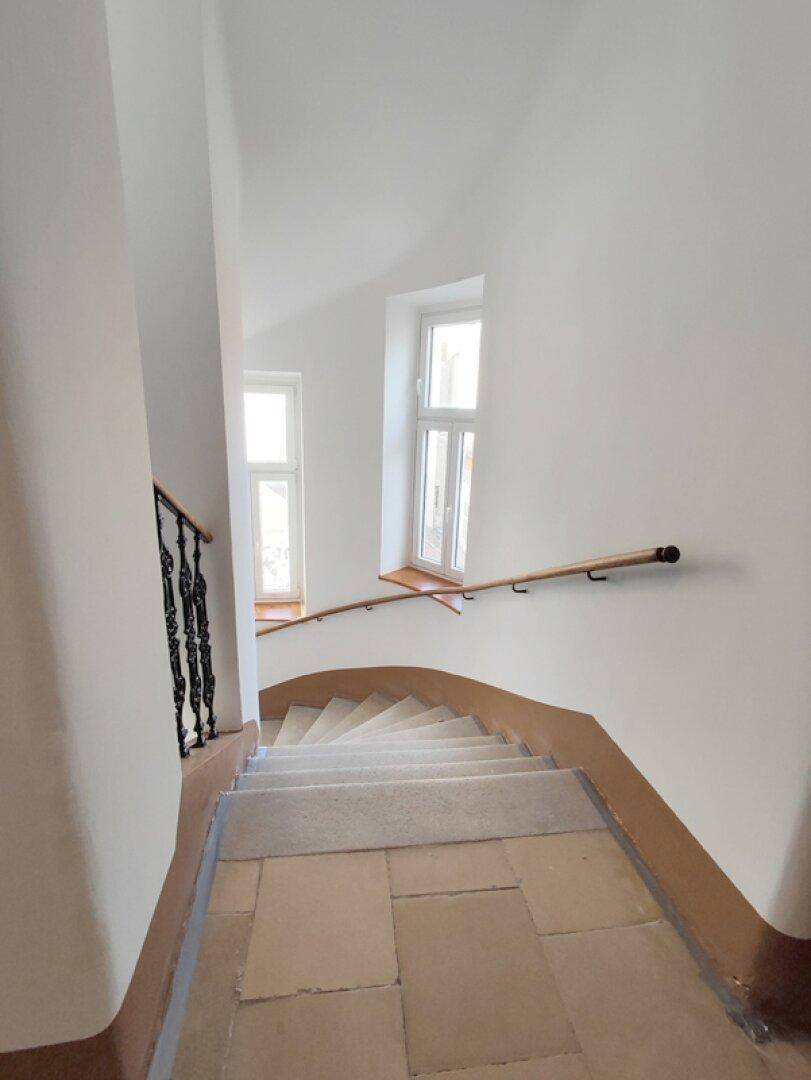 Breites Treppenhaus mit Steinstufen und einem schlichten Geländer, das nach unten führt.