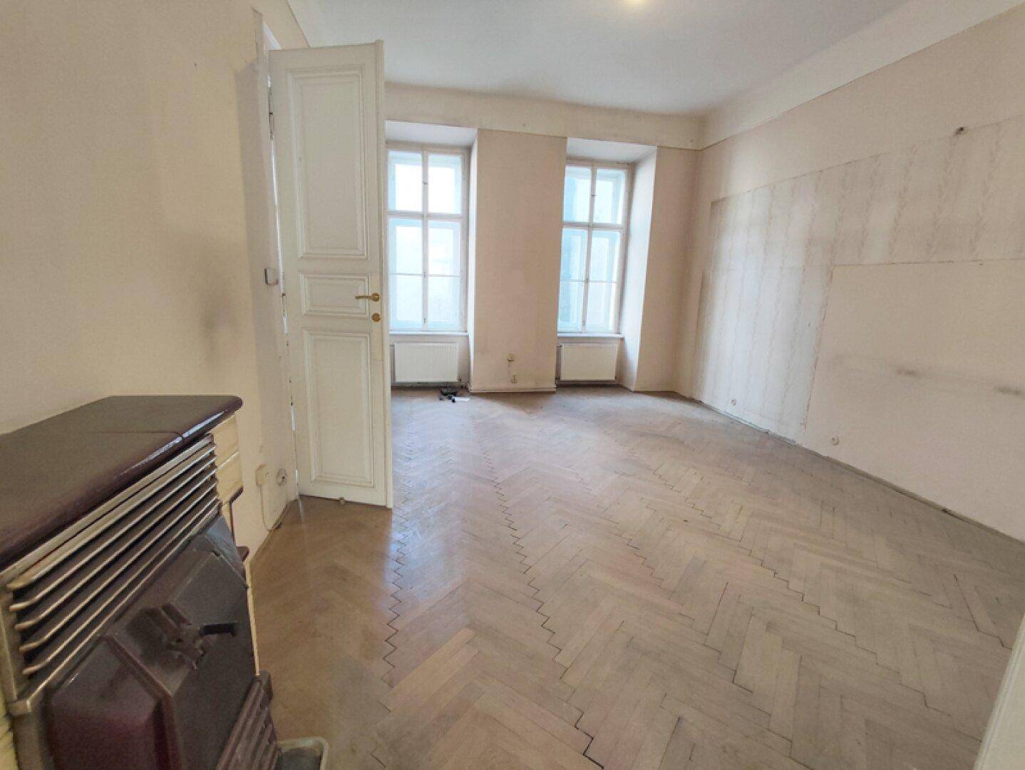 Geräumiges Wohnzimmer mit Fischgrätparkettboden, hohen Fenstern und einem traditionellen Ofen.