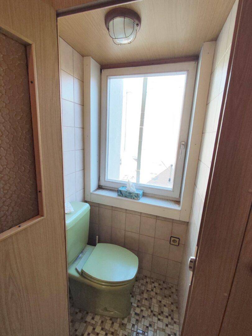 Kleines WC mit grünem WC, Fenster und gemusterten Bodenfliesen, renovierungsbedürftig.